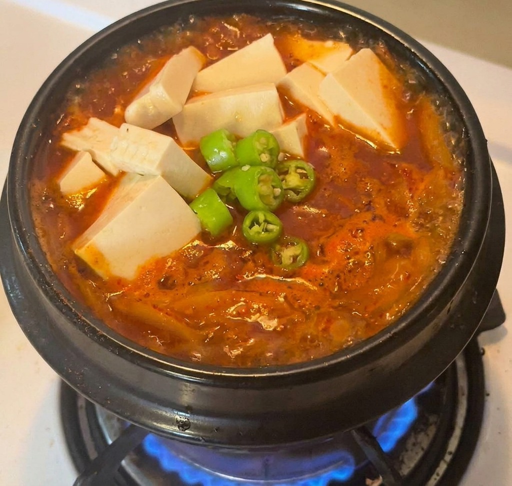 Un pot en pierre noire rempli de ragoût rouge, surmonté de cubes de tofu et de piments verts émincés au centre.