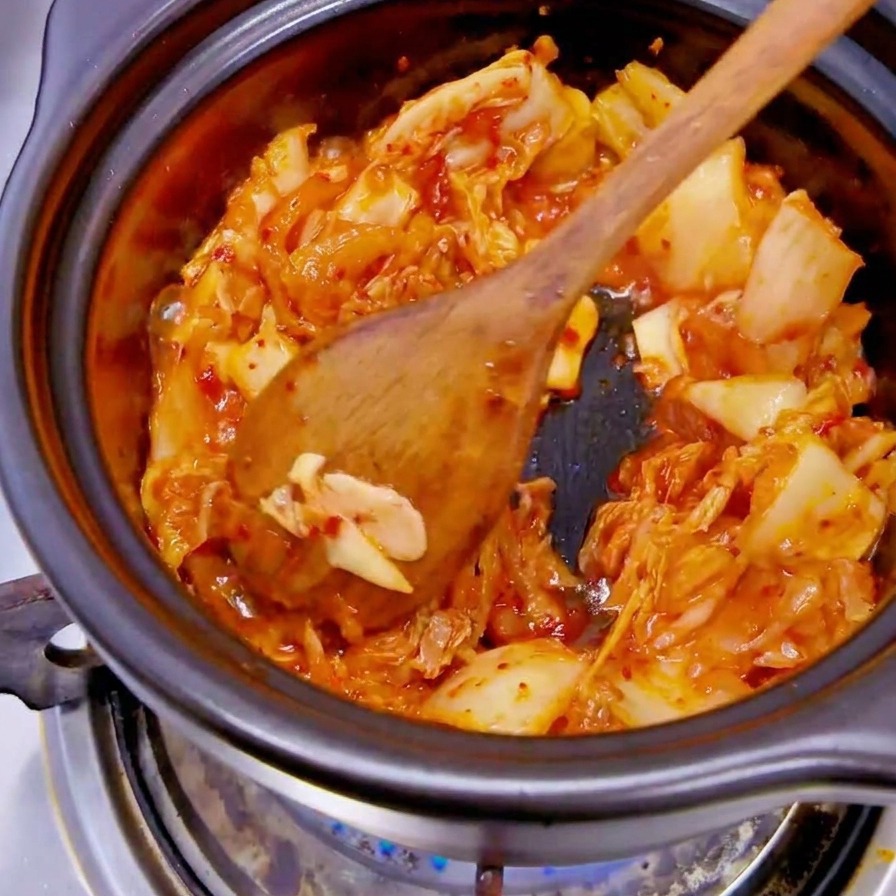 Une main déposant des tranches doignon blanc dans la marmite contenant le kimchi rouge.