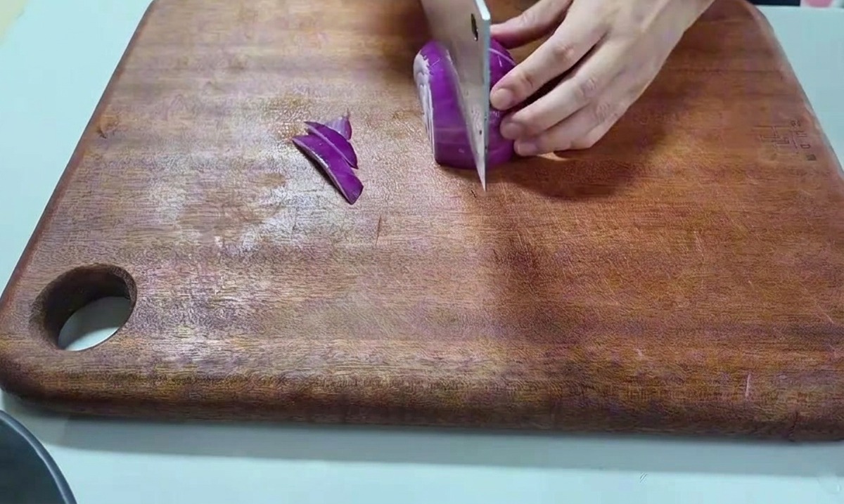 Une main tenant un oignon rouge et le coupant avec un grand couteau de cuisine sur une planche en bois.