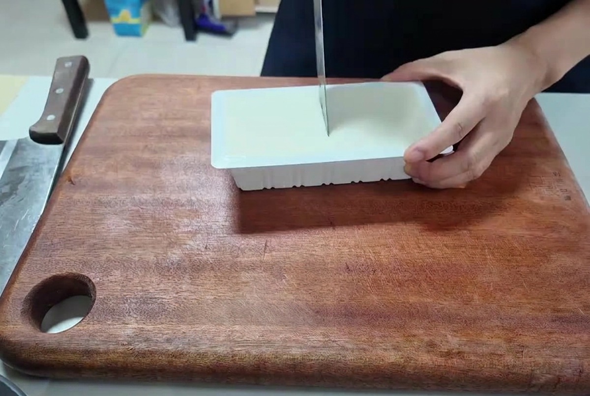 Une personne utilisant un couteau de cuisine pour couper un bloc rectangulaire de tofu blanc en petits cubes.