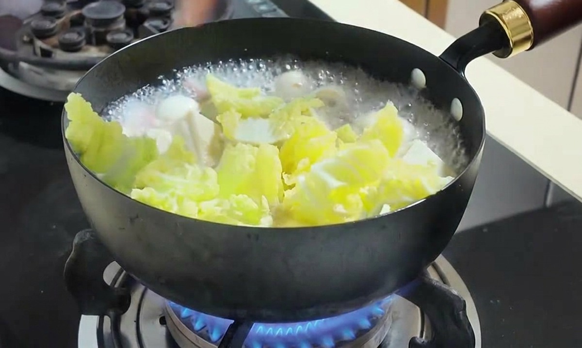 Une main ajoutant des grains de maïs jaune vif et des feuilles de chou vert dans une casserole de soupe bouillante.