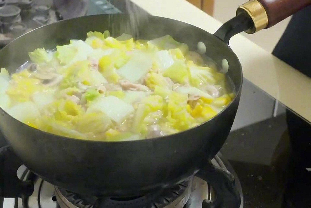 Une main saupoudrant du sel fin dans une poêle fumante remplie de légumes, de tofu et de soupe au thon.