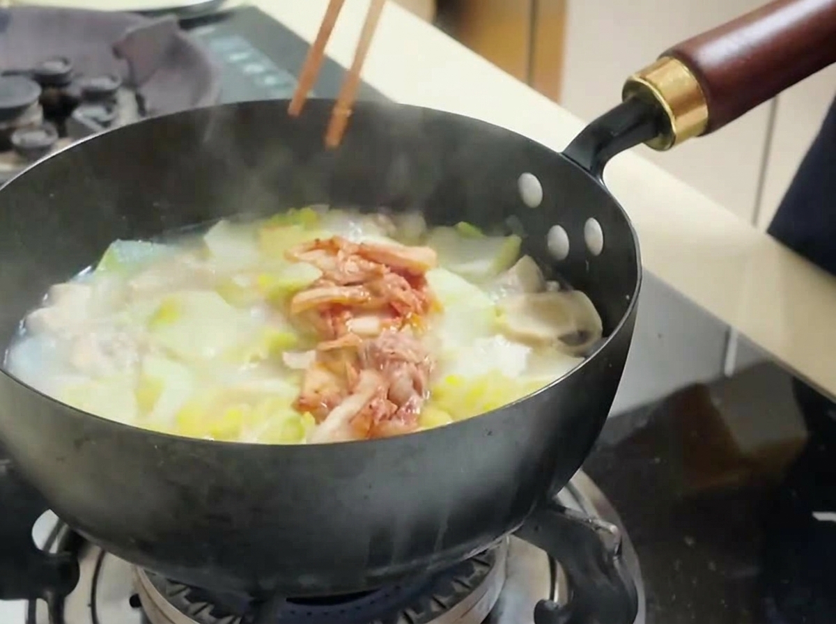 Une personne utilisant des baguettes pour ajouter du kimchi rouge haché dans une soupe frémissante.