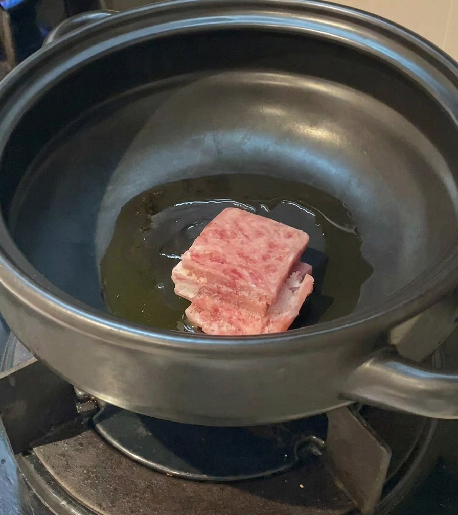 Des tranches de poitrine de porc crues et persillées déposées dans une marmite en terre cuite avec de lhuile chaude.