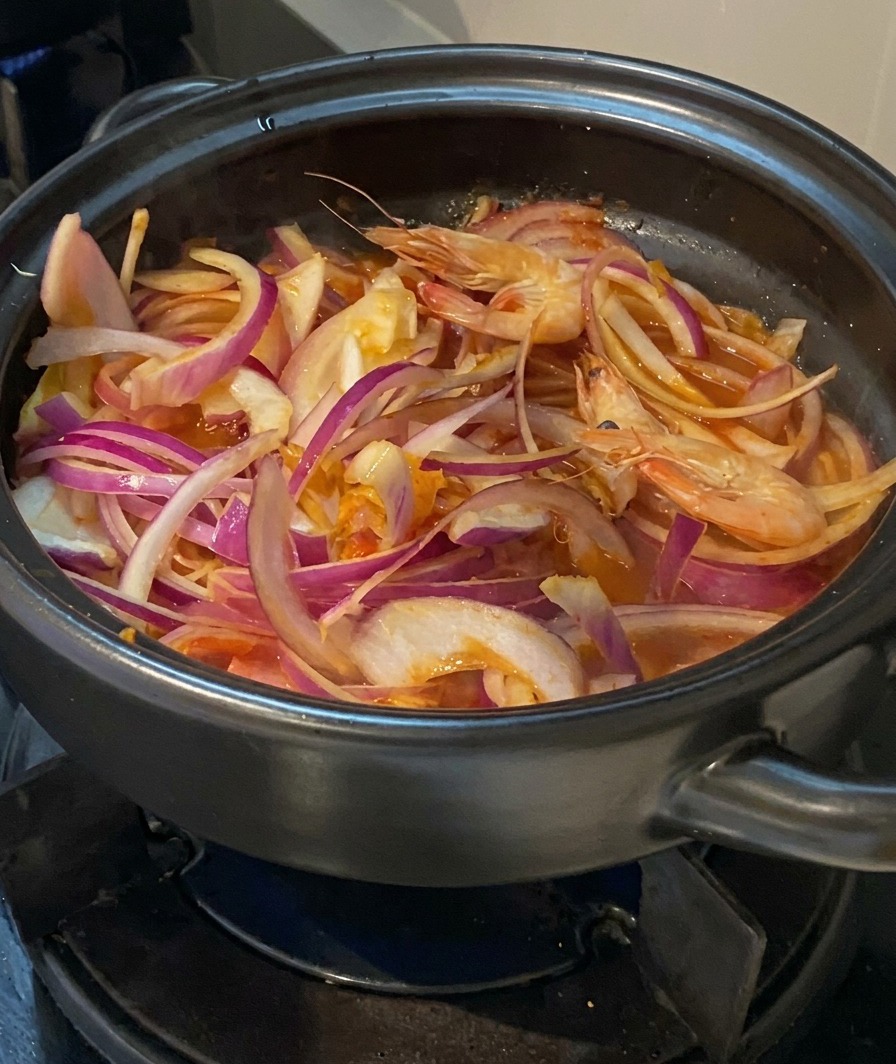 Des crevettes entières crues avec leurs têtes ajoutées dans une marmite remplie doignons, de kimchi et de porc.