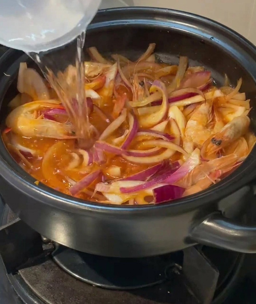 De leau claire versée depuis un récipient dans une marmite en terre cuite garnie de crevettes, doignons et de kimchi.
