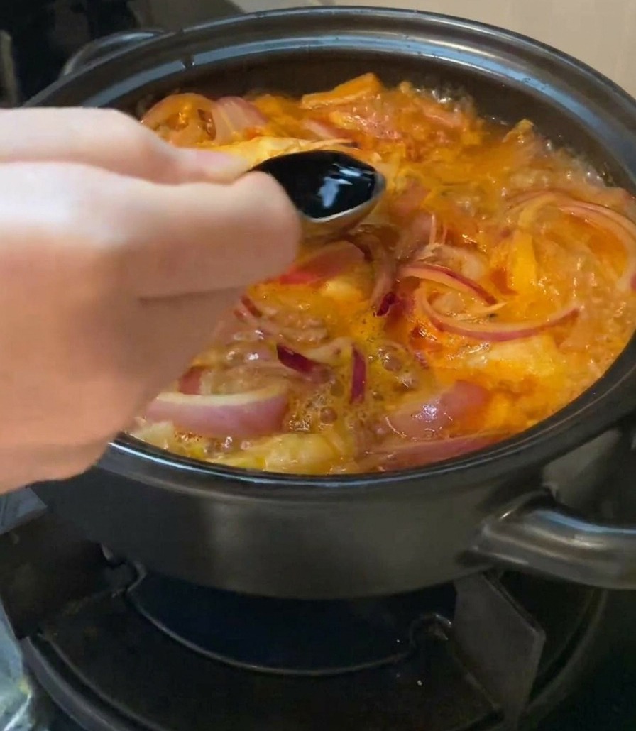 Une main tenant une cuillère en métal et versant de la sauce soja claire dans une marmite frémissante de soupe au kimchi et aux oignons rouges.