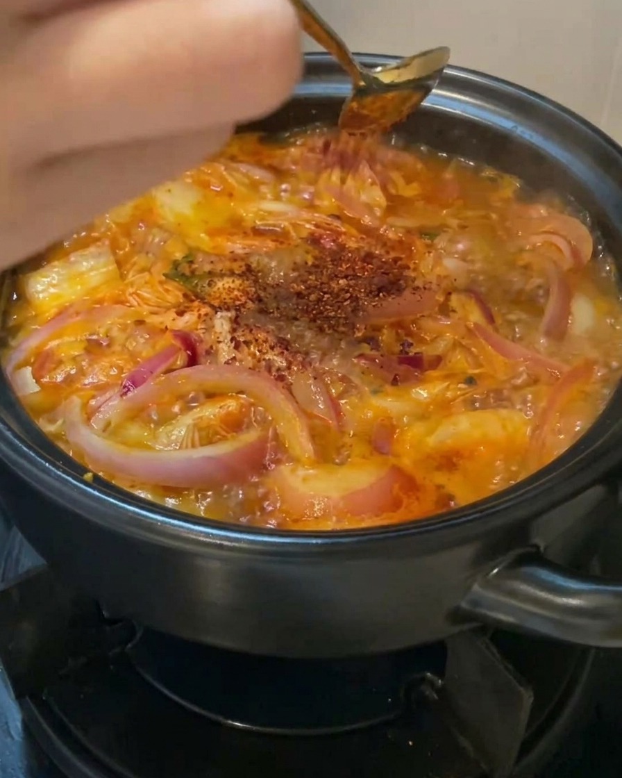 Une main saupoudrant de la poudre de piment rouge vif avec une cuillère dorée au-dessus dune soupe frémissante au kimchi et aux oignons.