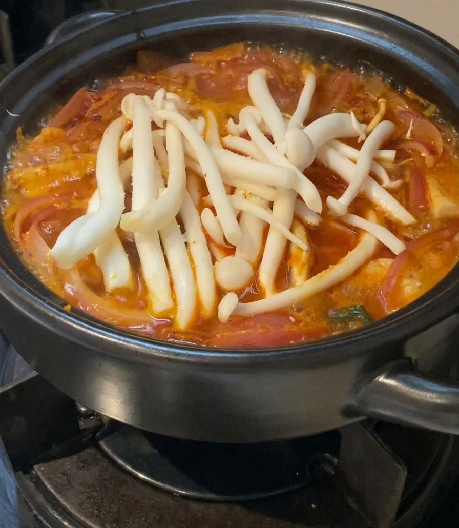 Une grappe de champignons blancs frais (shimeji) reposant à la surface dune soupe de kimchi rouge frémissante dans une marmite en céramique noire.