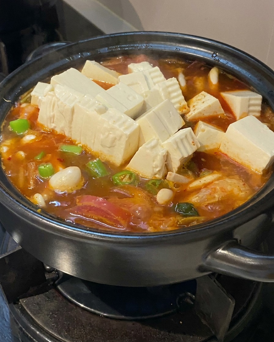Plusieurs cubes de tofu blanc frais et des rondelles de piments verts ajoutés à une soupe frémissante rouge orangé dans une marmite noire.