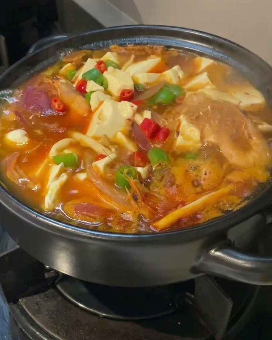 Un plan rapproché de la soupe au kimchi et tofu mijotant dans une marmite noire, garnie de rondelles de piments rouges et verts éclatants sur des cubes de tofu blanc.