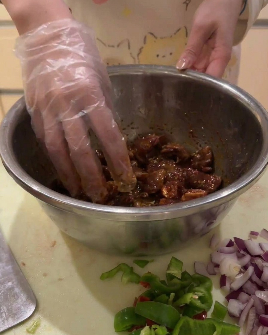 Main gantée mélangeant les cubes de bœuf marinés dans un bol en argent pour un enrobage uniforme.