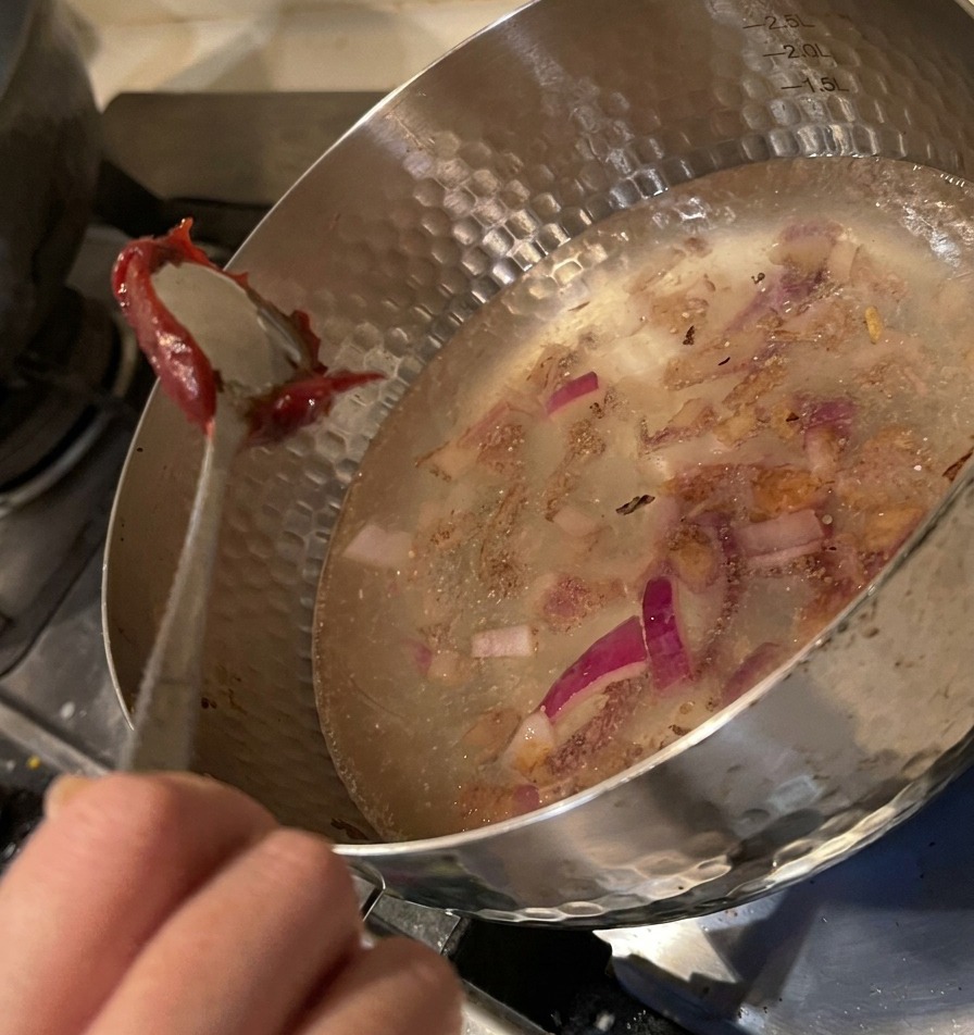 Cuillère déposant une dose de pâte de piment rouge épaisse dans la marmite remplie deau et doignons.