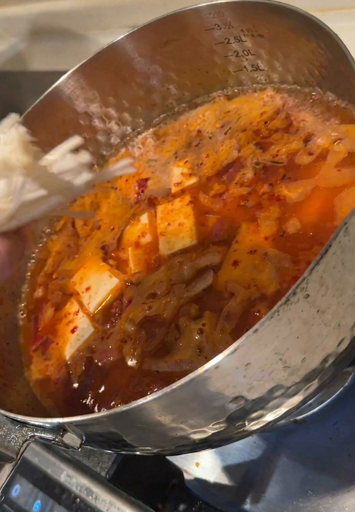 Main ajoutant un bouquet de champignons enoki dans une marmite de soupe kimchi bouillonnante contenant des cubes de tofu.