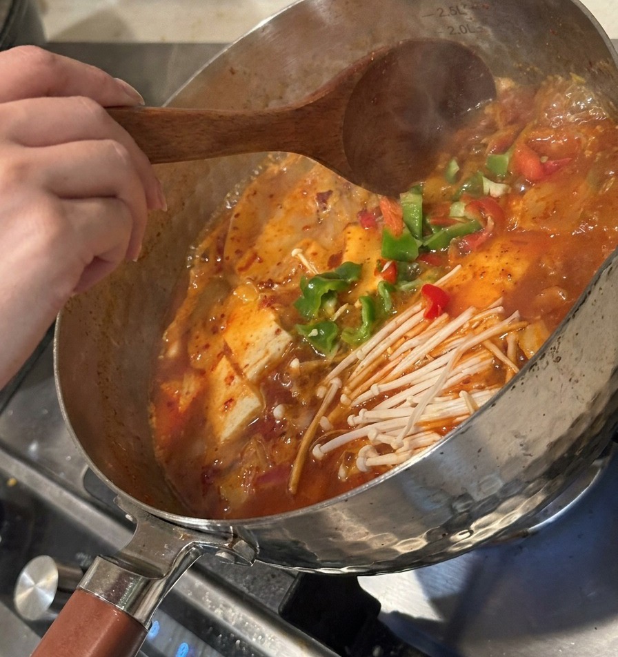 Poivrons verts émincés et kimchi fermenté ajoutés à la soupe bouillonnante et remués avec une cuillère en bois.
