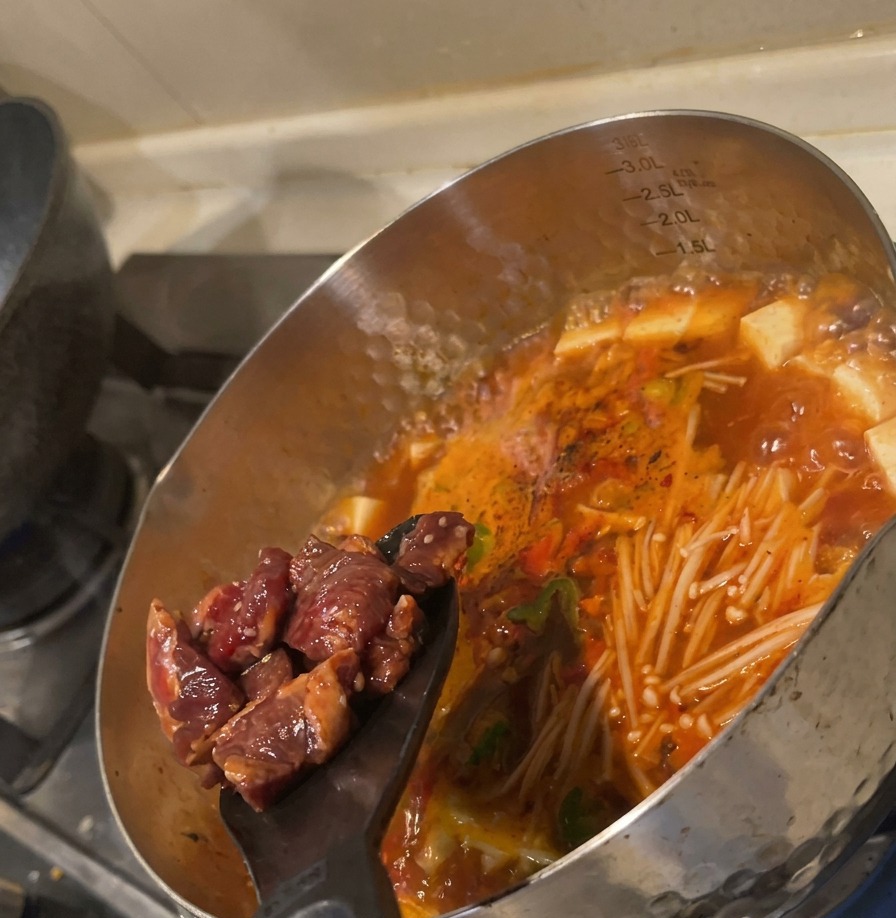 Cubes de bœuf crus marinés versés dune spatule noire dans la soupe rouge bouillante.