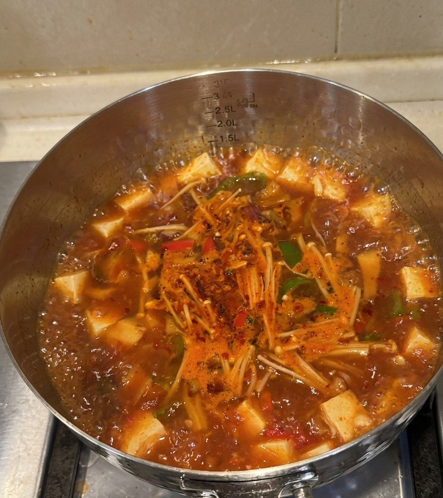 Marmite en inox remplie de soupe kimchi au bœuf bouillonnante, montrant la viande cuite, les enokis et les poivrons.