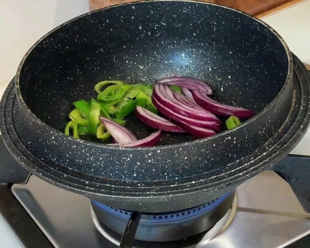 Tranches doignon rouge ajoutées aux oignons verts sautés dans un pot noir sur une flamme de gaz bleue.