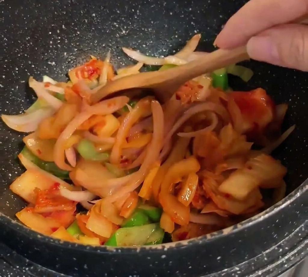 Une cuillère en bois mélangeant du kimchi orange-rouge vif à un mélange doignons sautés.