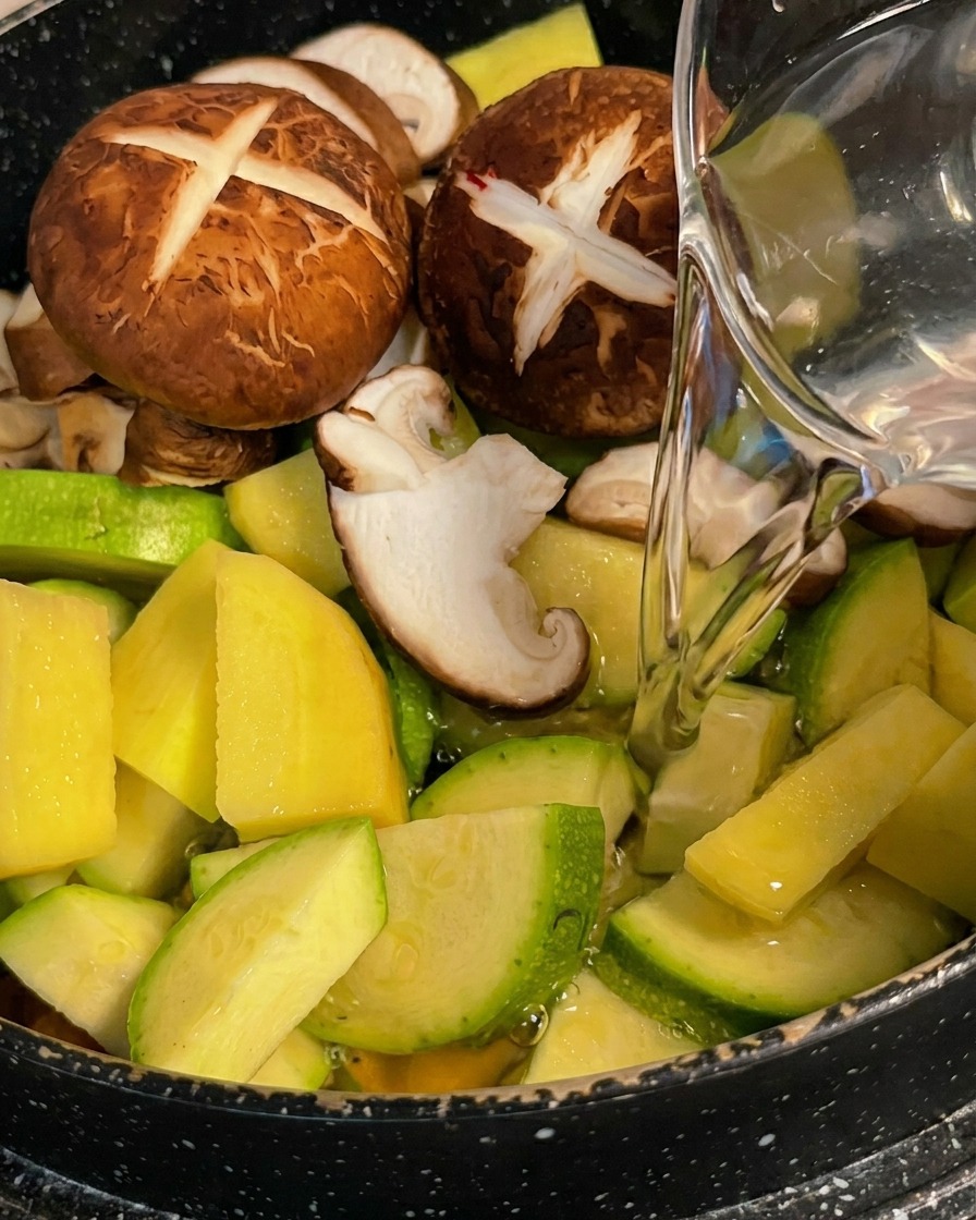 Eau claire versée dun pichet sur un mélange coloré de courgettes, pommes de terre et shiitakés dans un pot.