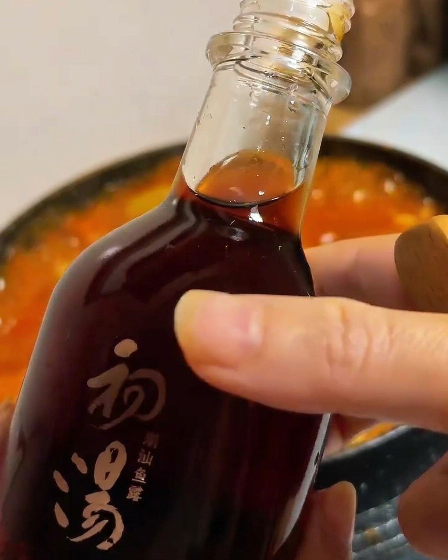 Une bouteille en verre sombre versant de la sauce poisson dans un pot de ragoût de tofu et légumes rouge.