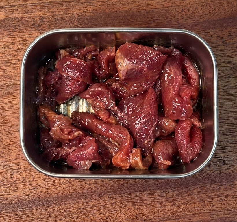 Tranches de bœuf cru marinées enrobées dune sauce sombre et brillante dans un récipient en inox.