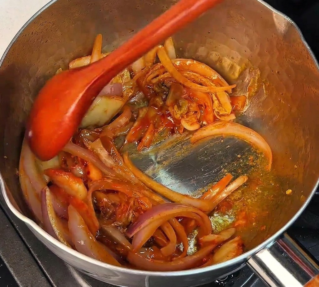 Une cuillère en bois mélangeant du kimchi haché et des oignons rouges dans une marmite chaude.