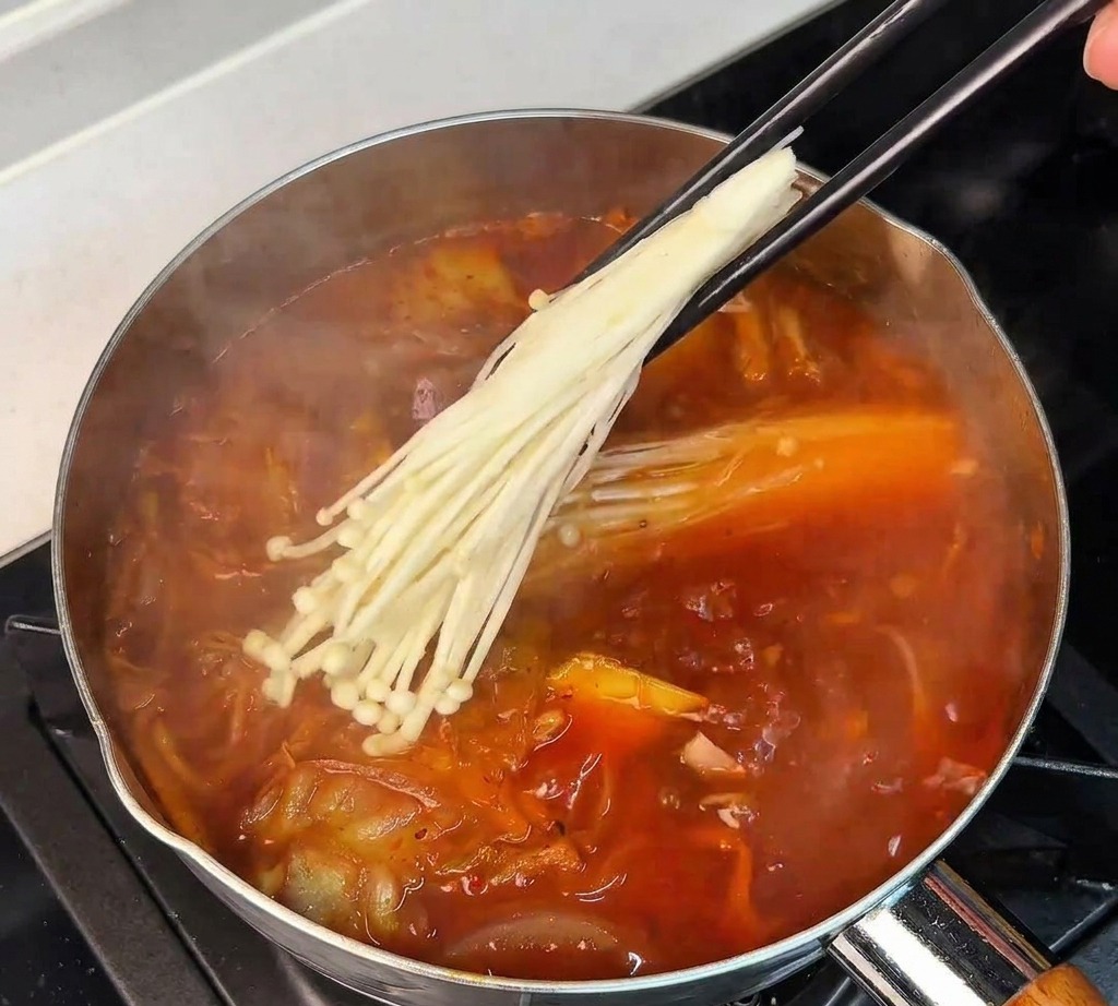 Des baguettes déposant un bouquet de champignons enoki blancs dans la soupe rouge frémissante.