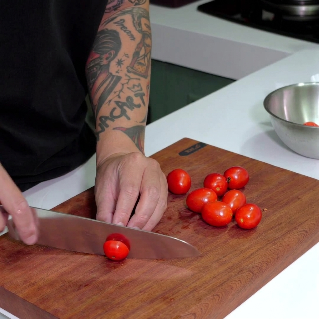 Un couteau de chef tranchant des tomates cerises rouge vif sur une planche en bois sombre.