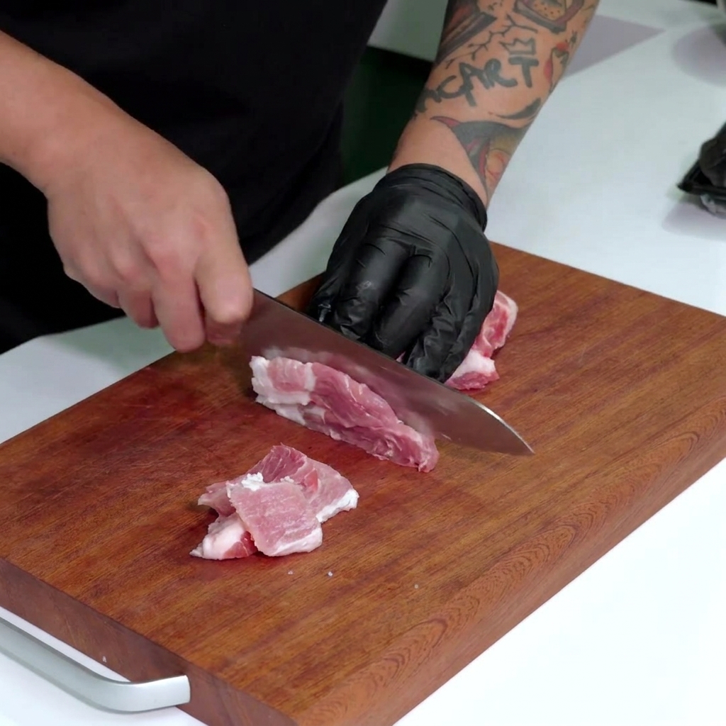Une main gantée utilisant un grand couteau pour couper de la poitrine de porc marbrée en fines lamelles sur une planche en bois.
