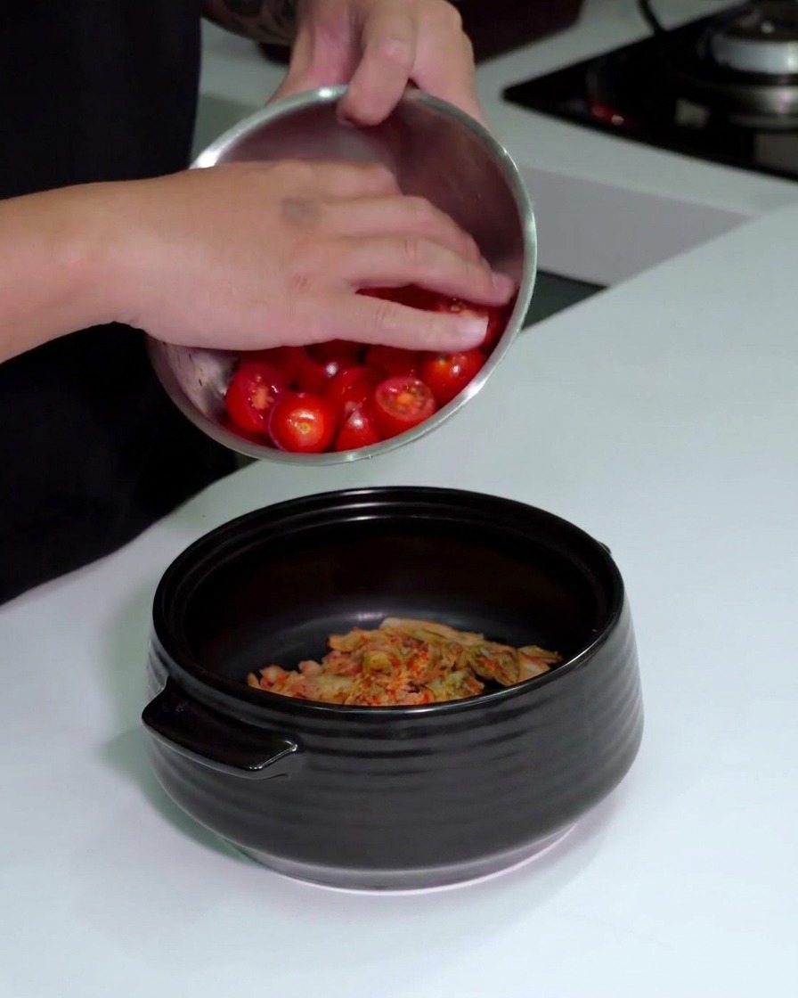 Une personne versant un bol de tomates cerises coupées dans une marmite contenant déjà le kimchi haché.