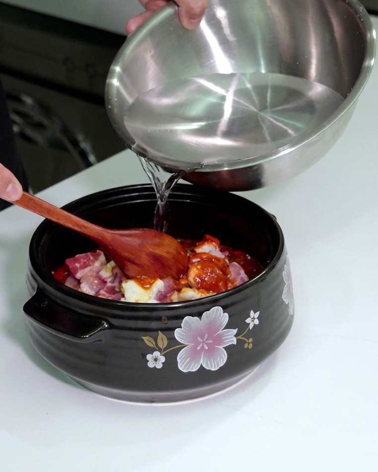 Une personne versant de leau dun bol mélangeur en inox dans une marmite contenant la viande et les légumes assaisonnés.