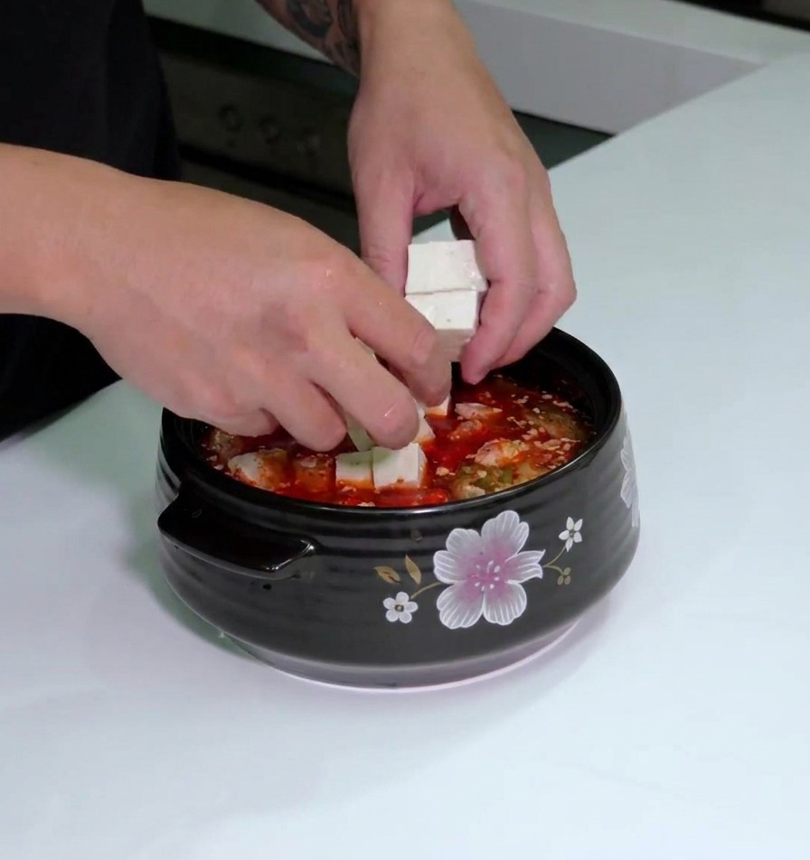 Des mains plaçant des dés de tofu frais dans une marmite en céramique remplie de soupe au kimchi et de poitrine de porc.