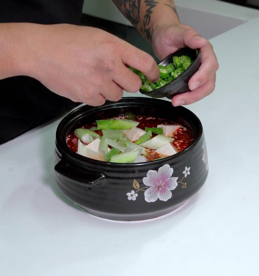 Des piments verts hachés saupoudrés depuis un petit bol sur le dessus dune marmite de soupe au kimchi avec tofu et poireaux.