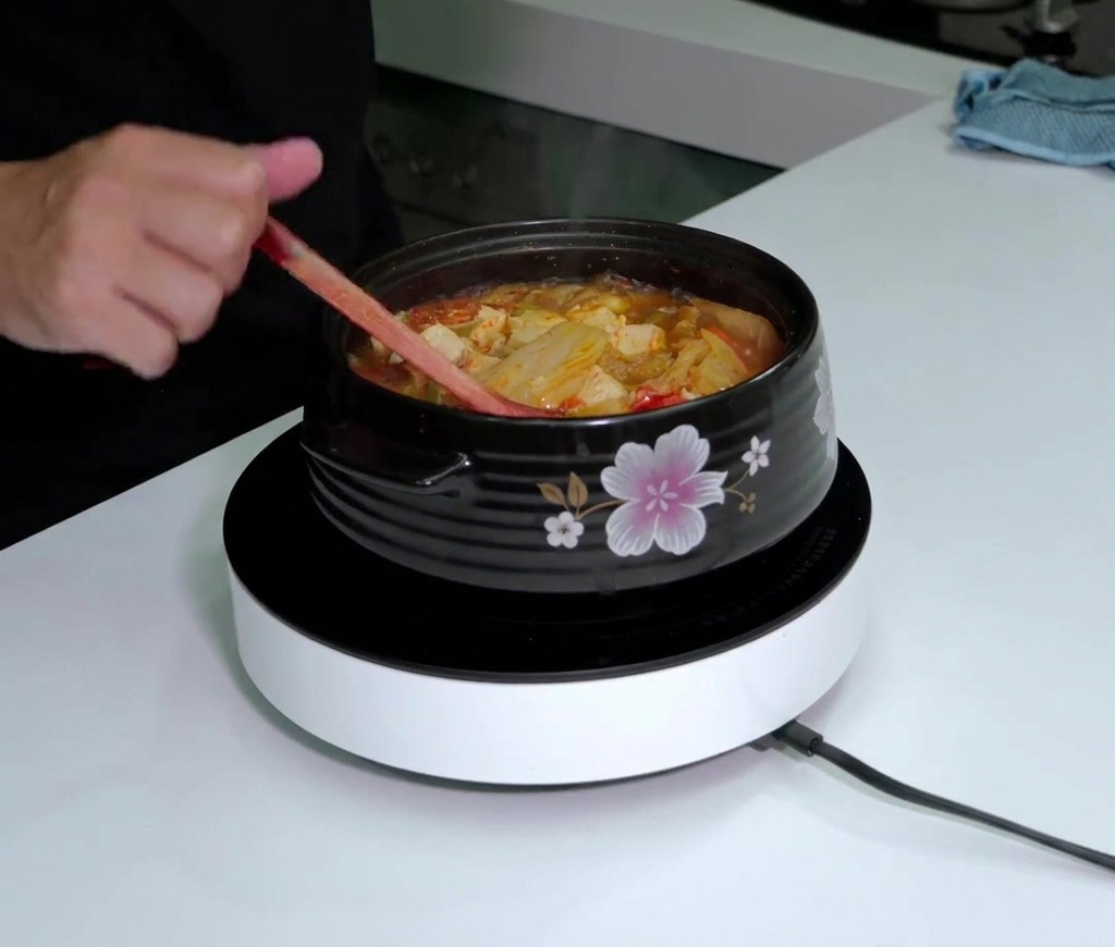 Une bouteille de sauce Tabasco tenue au-dessus dune marmite fumante de soupe au kimchi rouge pendant quon mélange avec une cuillère en bois.