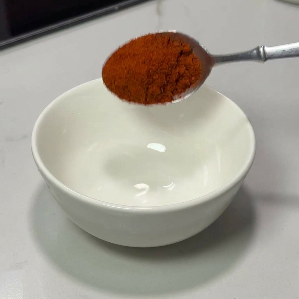 Une cuillère versant du piment rouge vif en poudre dans un bol en céramique blanc.