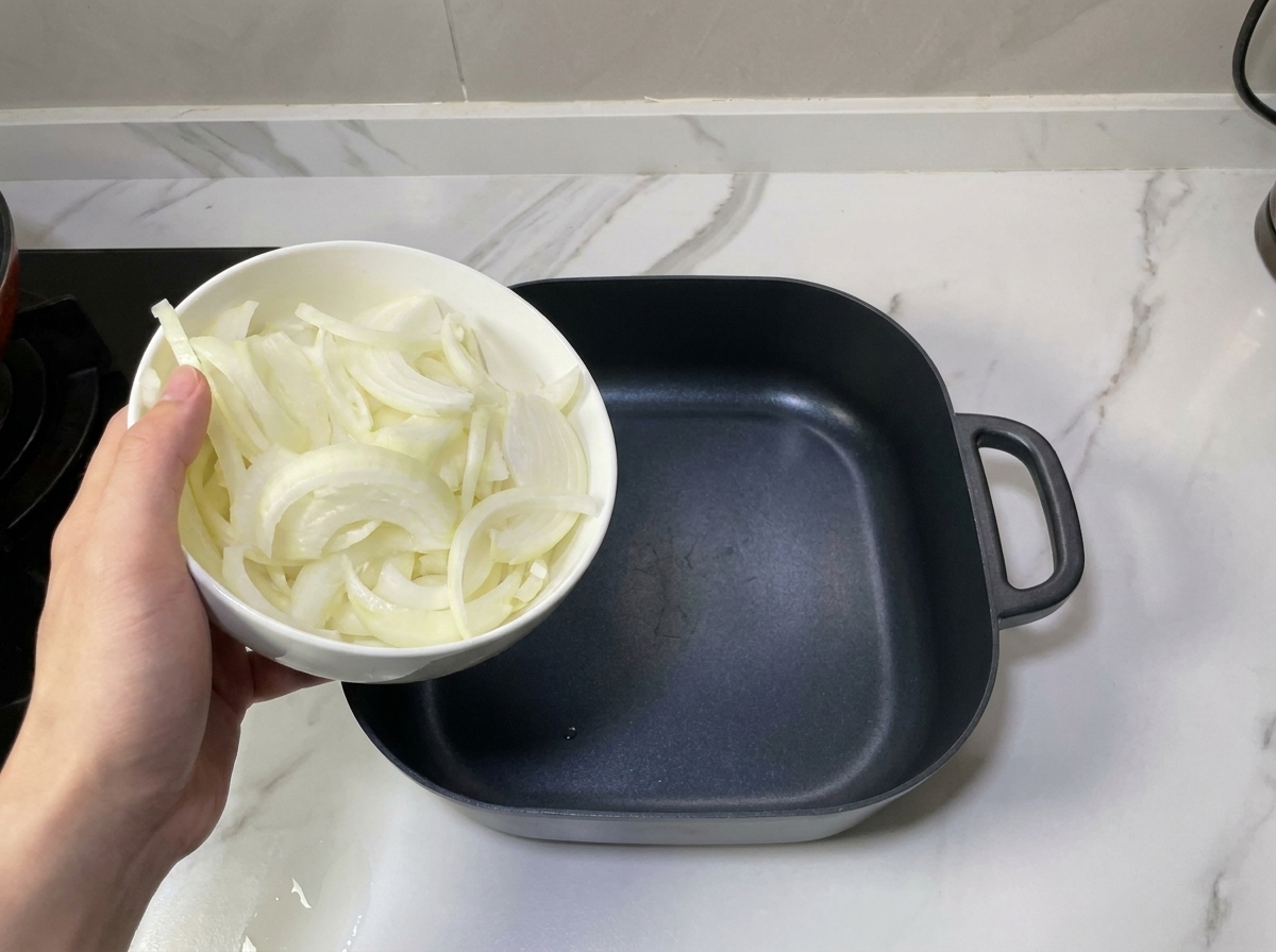 Une main déposant des oignons émincés au fond dune marmite noire vide pour créer une base aromatique.