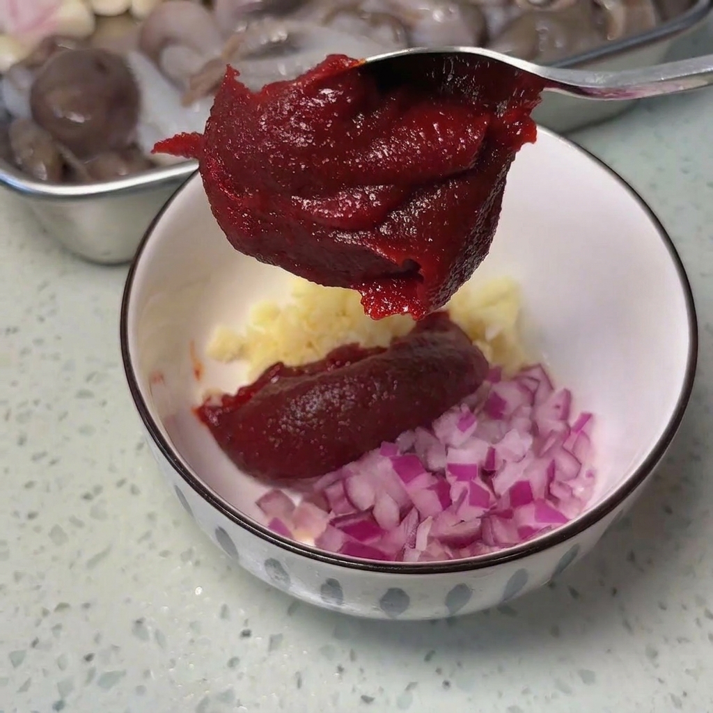 Une cuillère en métal déposant de la pâte de piment rouge épaisse dans un bol contenant de lail et de loignon rouge.