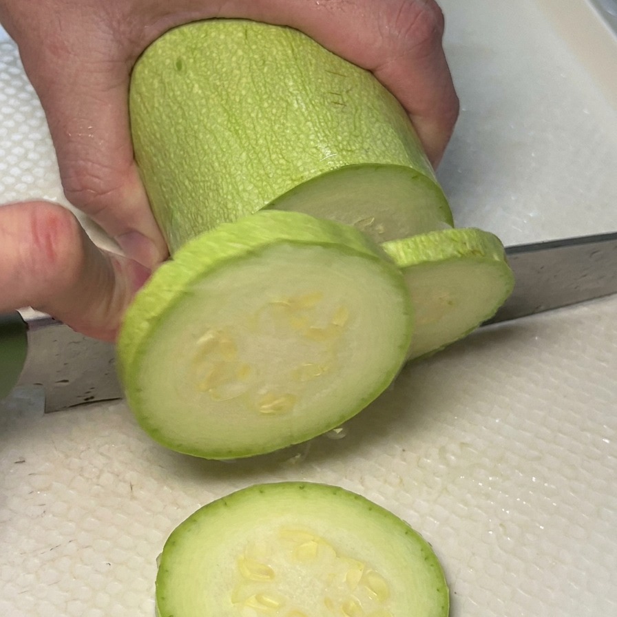 Mains découpant une courgette vert clair en rondelles sur une planche à découper.