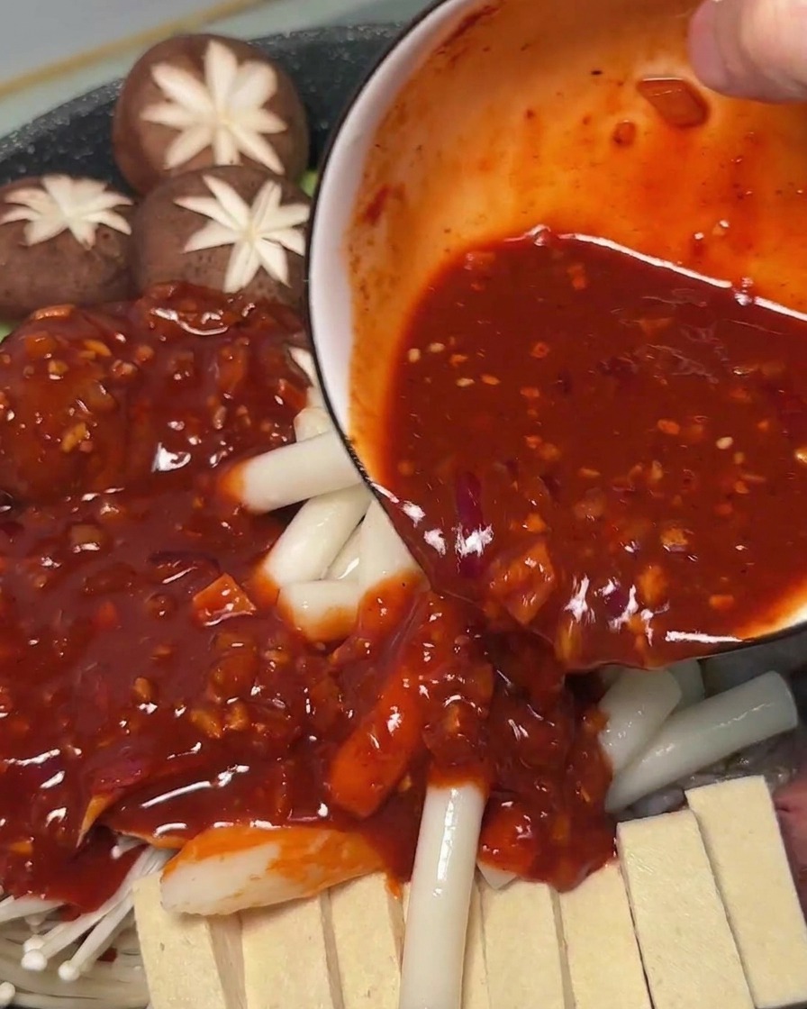 Sauce rouge épicée versée sur les ingrédients crus et les gâteaux de riz dans la marmite.