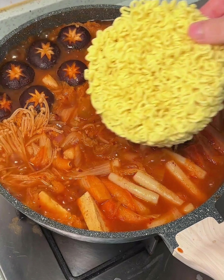 Une main déposant un bloc de nouilles ramen sèches dans le bouillon rouge bouillonnant.
