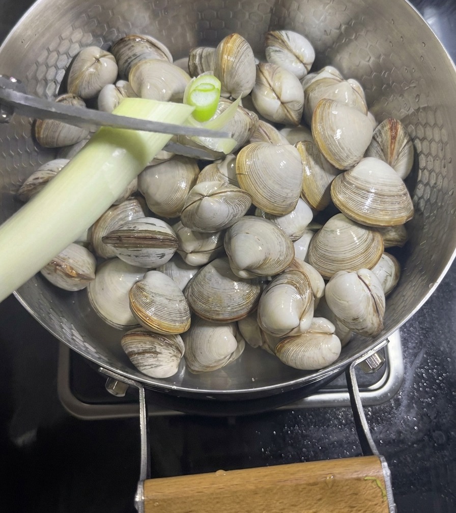 Une main utilisant des ciseaux pour couper des oignons verts au-dessus dune poêle remplie de palourdes et de tranches de gingembre.