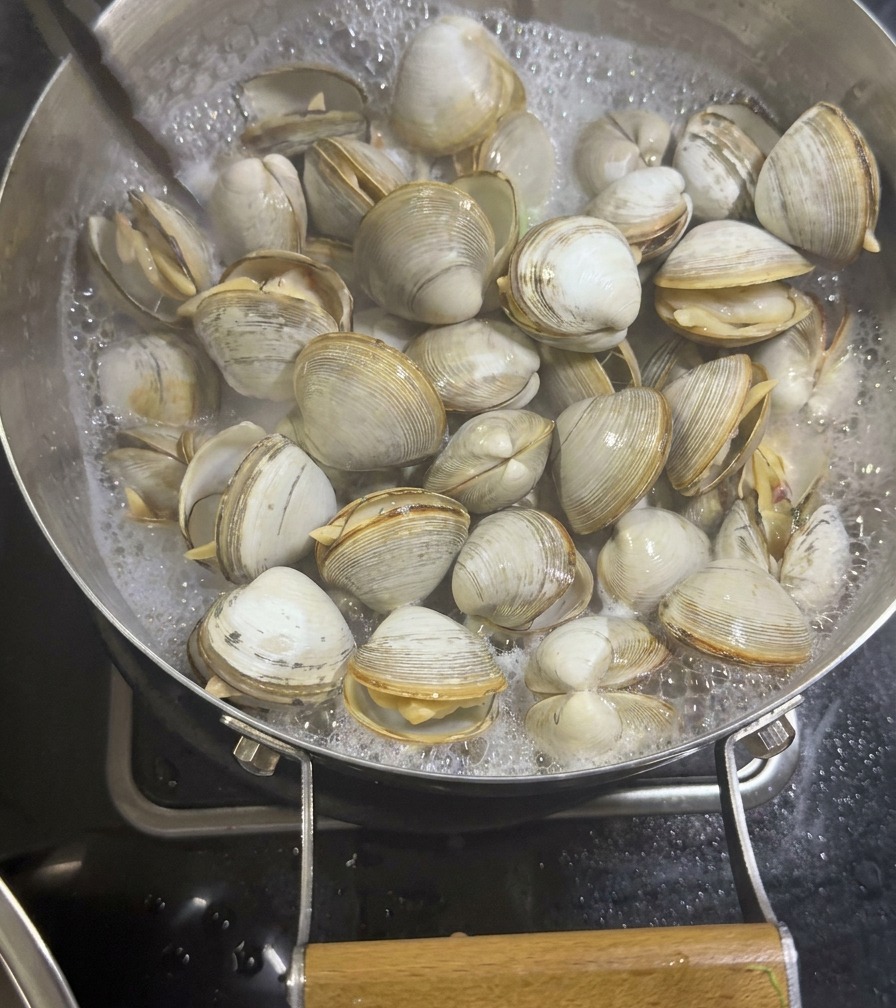 Une pince métallique soulevant des palourdes blanches ouvertes hors dune poêle contenant un bouillon de mer laiteux.