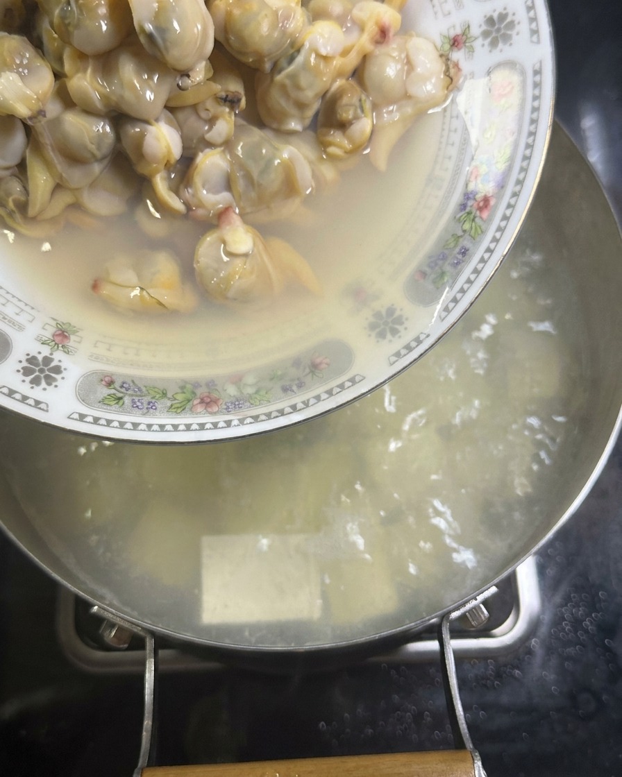 Une assiette de chair de palourdes cuites versée dans une casserole de bouillon blanc frémissant contenant des dés de tofu.