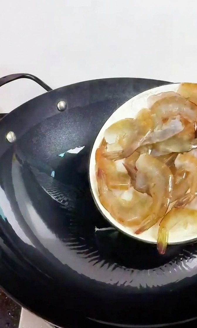 Une main tenant une assiette blanche et versant des crevettes fraîches translucides dans un wok en fonte préchauffé.