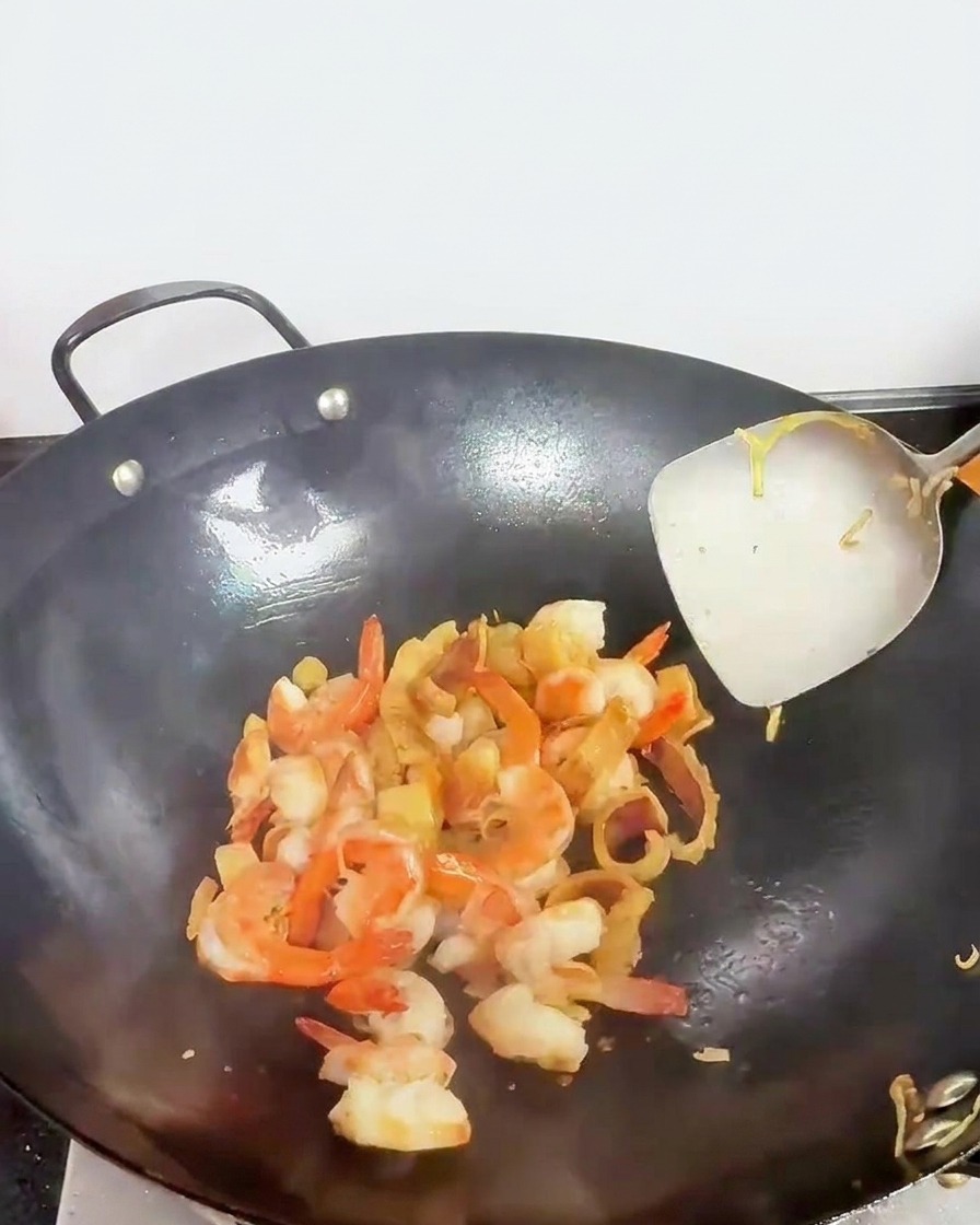 Des morceaux de fruits de mer, dont des crevettes rosies, remués dans un wok avec une spatule métallique.