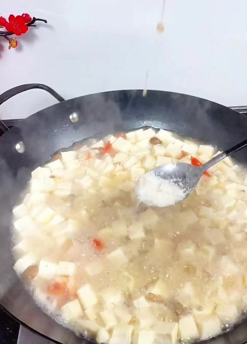 Une cuillère métallique versant du sel fin dans un grand wok rempli de dés de tofu et fruits de mer mijotant.