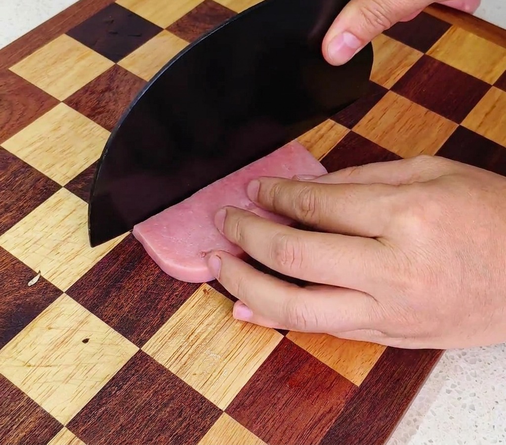 Une personne utilisant un grand couteau de chef noir pour couper une saucisse de jambon rose en dés sur une planche à découper en bois.