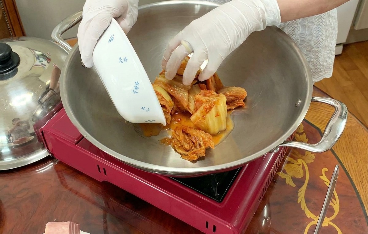 Une personne avec des gants blancs dépose du kimchi émincé depuis un bol fleuri au centre dune grande marmite argentée.