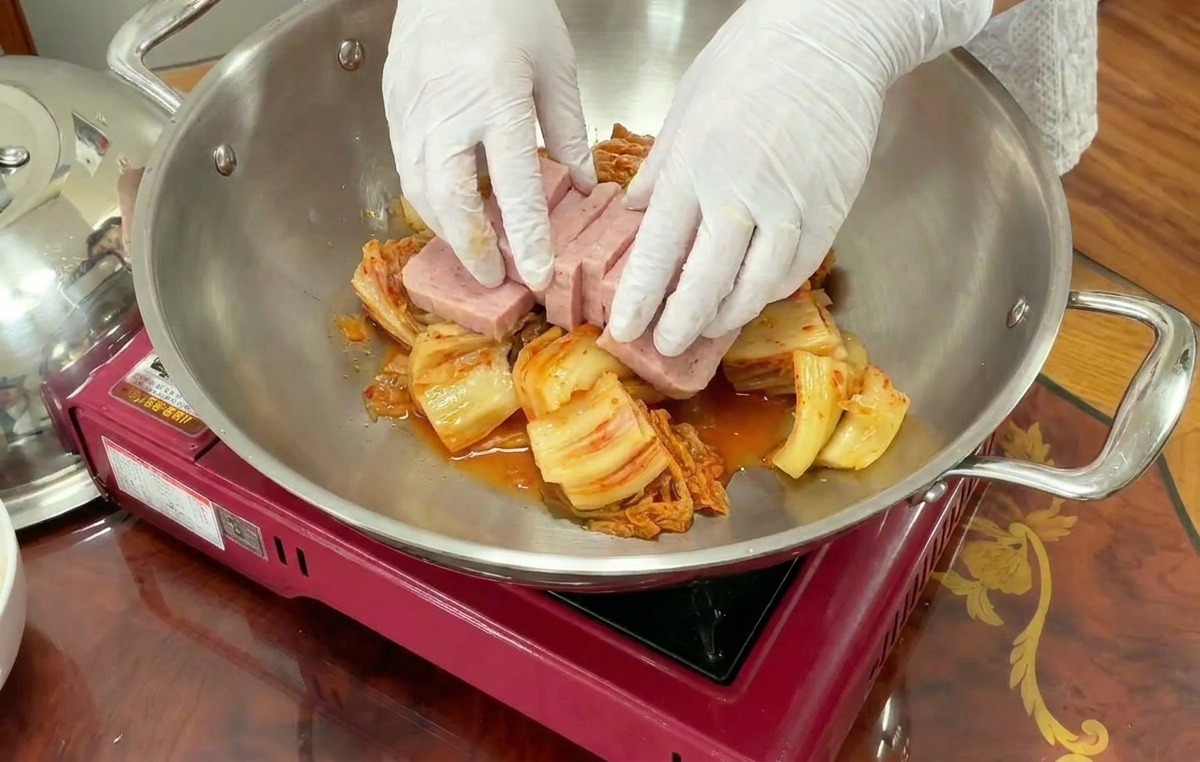 Des mains gantées disposent soigneusement des tranches rectangulaires de jambon rose sur un lit de kimchi dans un wok.